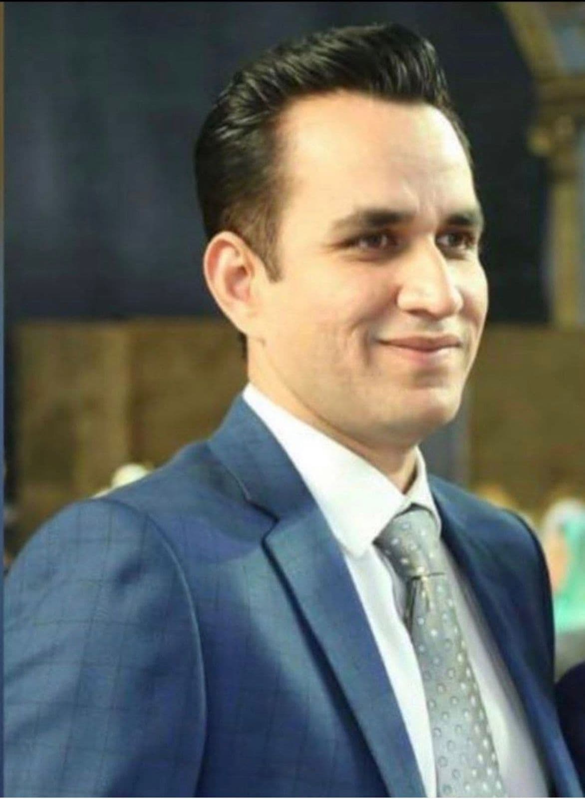 Smiling man with dark hair in a blue suit and grey patterned necktie.